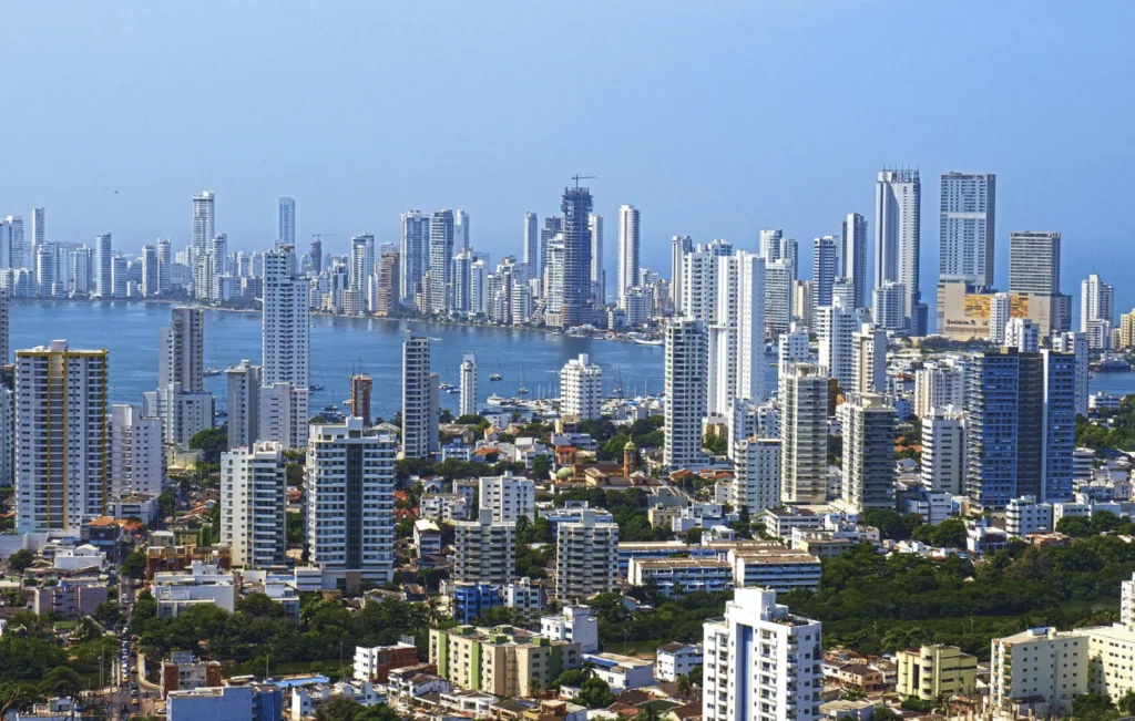 Panoramic aerial view of Cartagena's modern skyline featuring gleaming white skyscrapers and residential buildings along the Caribbean coast, with the historic district visible in the foreground and turquoise waters stretching to the horizon.