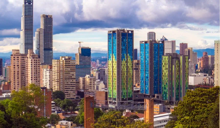 A cityscape featuring tall modern buildings, including two blue and green luxury high-rises, with trees in the foreground and a calm, cloudy sky overhead.