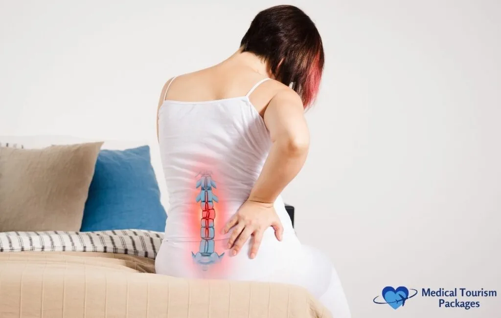 Woman in white tank top experiencing lower back pain, shown from behind with illuminated red overlay on spine, sitting on beige couch with blue pillow, illustrating common symptom leading to breast reduction surgery