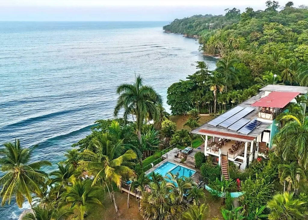 Una moderna casa con paneles solares y piscina rodeada de palmeras, con vistas al océano y a la exuberante costa panameña, perfecta para lujosos retiros de bienestar o yoga.