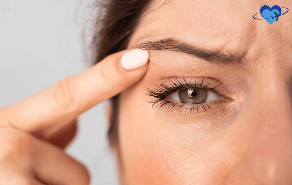 Close-up of a person's eye with a finger gently pulling the outer corner of the eyebrow, showcasing an eye lift effect. A heart with an airplane icon in the top right corner hints at a journey to Colombia for blepharoplasty.