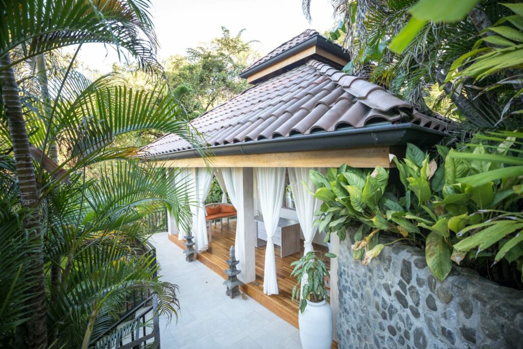 Un pequeño pabellón con techo de tejas y cortinas blancas está rodeado de plantas tropicales y muros de piedra, con suelos de madera y asientos al aire libre, perfecto para retiros de bienestar en Costa Rica.