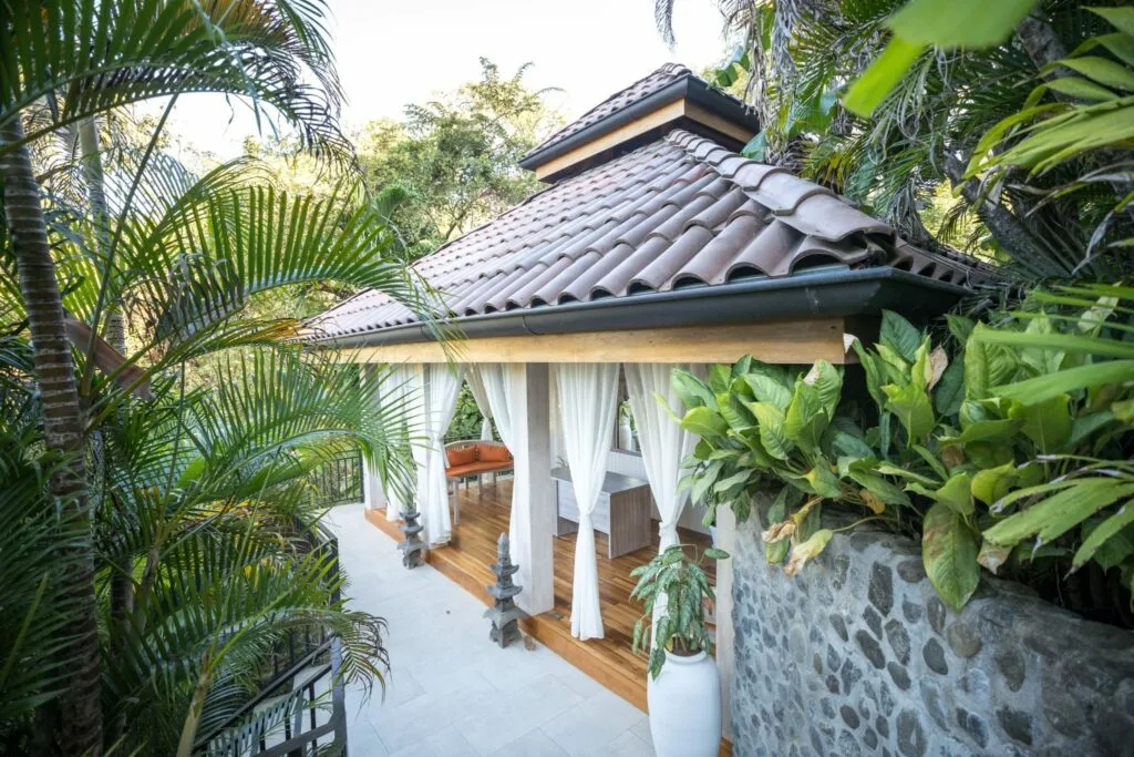 A small pavilion with a tiled roof and white curtains is surrounded by tropical plants and stone walls, featuring wooden floors and outdoor seating—perfect for wellness retreats in Costa Rica.