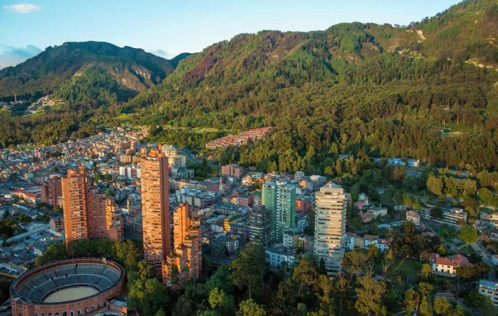 Aerial panorama of Bogotá's Macarena neighborhood at sunset, featuring tall terracotta-colored residential towers, a circular bullring, and lush green Eastern Hills (Cerros Orientales) rising dramatically in the background under golden evening light