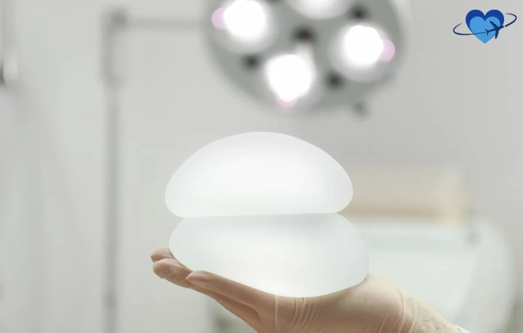 Close-up photograph of a gloved hand holding a smooth, round silicone breast implant in a sterile medical environment, with surgical lights visible in the blurred background, demonstrating the translucent and natural appearance of the implant.