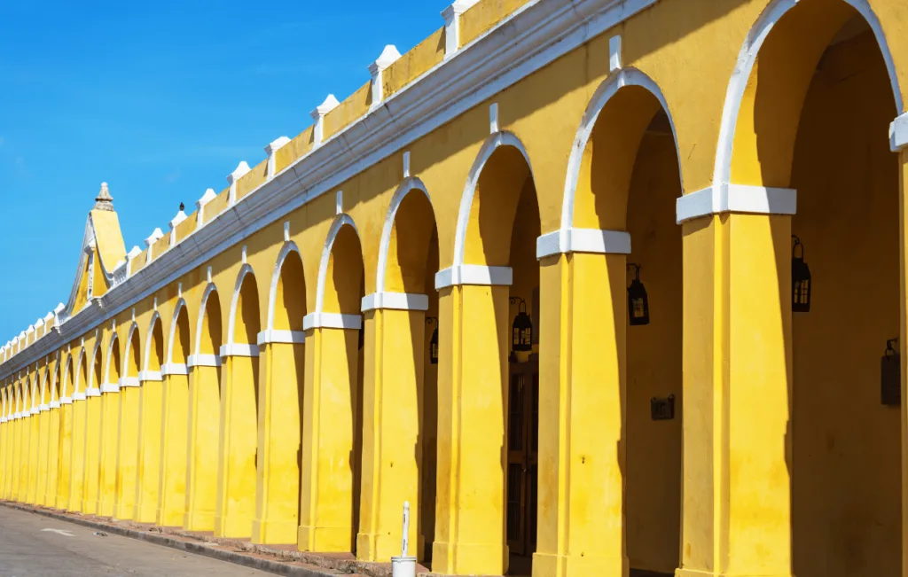 Historic yellow colonial building featuring a long series of pristine white archways and lanterns, displaying classical Spanish colonial architecture against vivid blue sky, part of Las Bóvedas in Cartagena's Walled City.