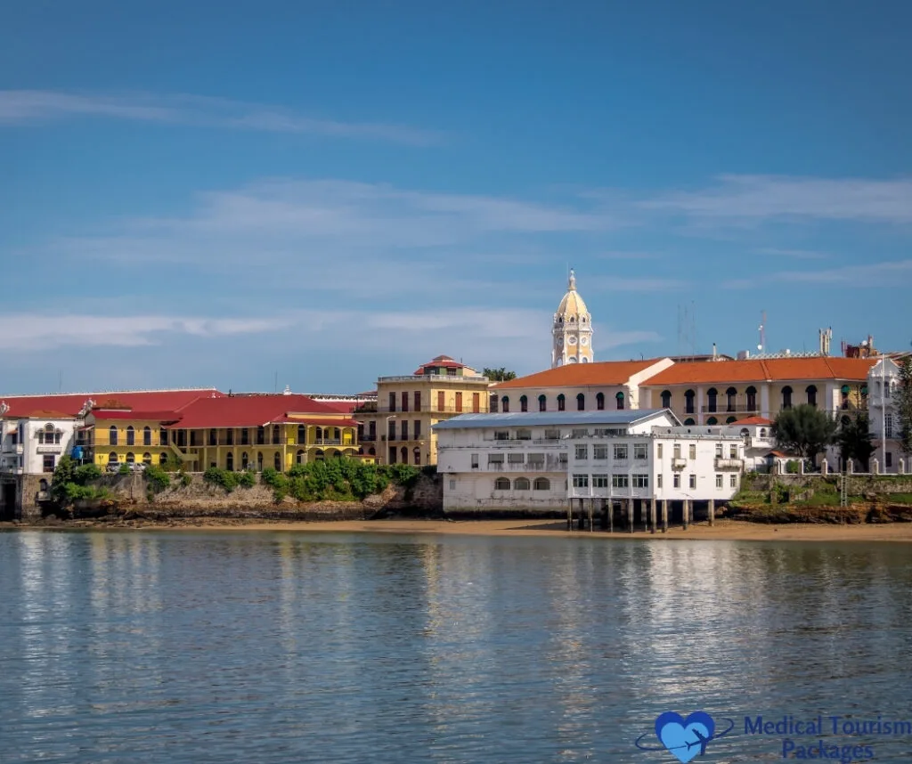 Vista frente al mar de coloridos edificios coloniales con tejados rojos y una torre del reloj bajo un cielo azul, reflejado en aguas tranquilas: una escena impresionante que muestra las principales atracciones turísticas que ofrece Panamá para vivir experiencias turísticas inolvidables.