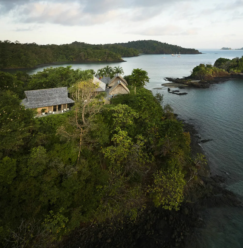 Vista aérea de una casa costera rodeada de densa vegetación, con aguas tranquilas y barcos visibles a lo lejos bajo un cielo parcialmente nublado.