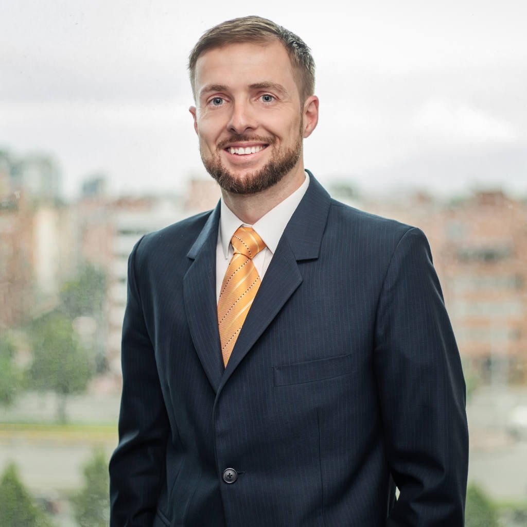 craig dempsey A man in a dark suit and orange tie stands indoors in front of a window with an urban cityscape in the background.