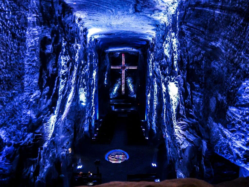An underground church hall with blue-lit stone walls exudes a calm atmosphere, featuring a large illuminated cross at the end of the aisle for a touch of serene luxury.