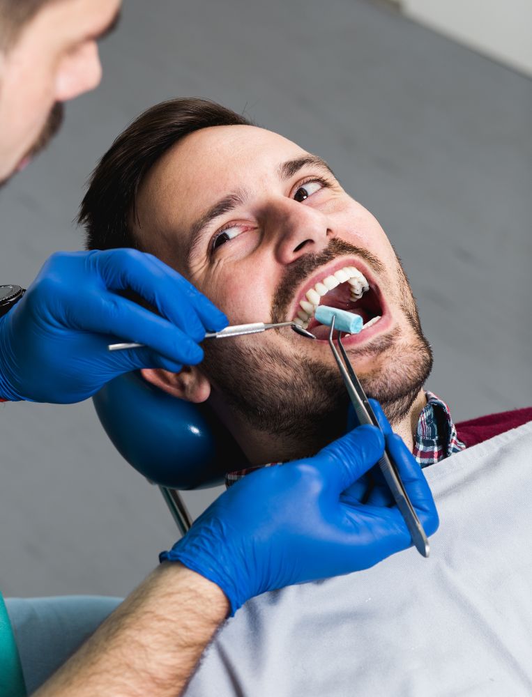 dental A dentist wearing blue gloves examines a man's teeth using dental tools while the man sits in a dental chair, highlighting affordable dental implants available in Latin America.