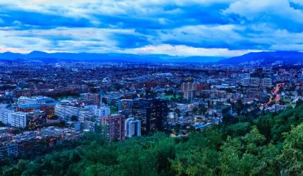 A wide view of a cityscape at dusk, where luxury buildings rise among calm, green foreground trees, set against mountains and a cloudy sky.