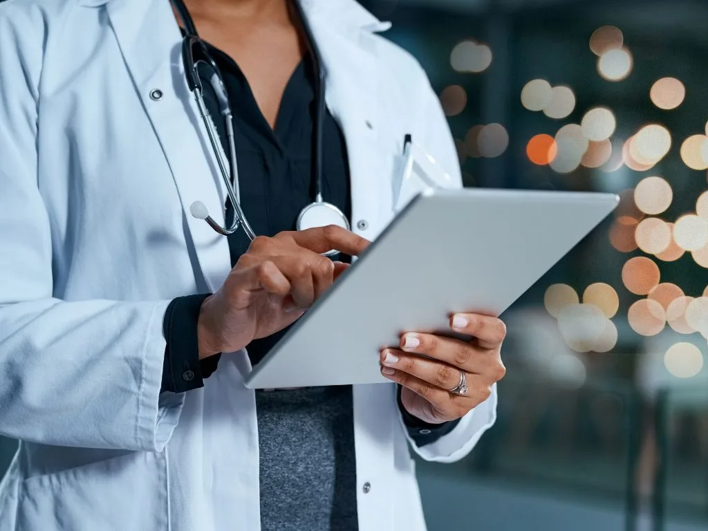 A person in a white lab coat and stethoscope calmly uses a tablet device, with blurred luxury lights glowing softly in the background.