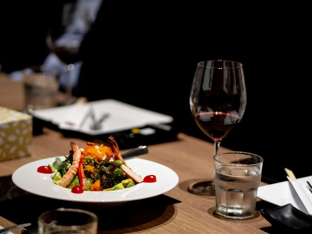 A plate of salad with shrimp and garnish sits on a wooden table next to a glass of red wine, a glass of water, and dining utensils, creating a calm atmosphere with a touch of luxury.