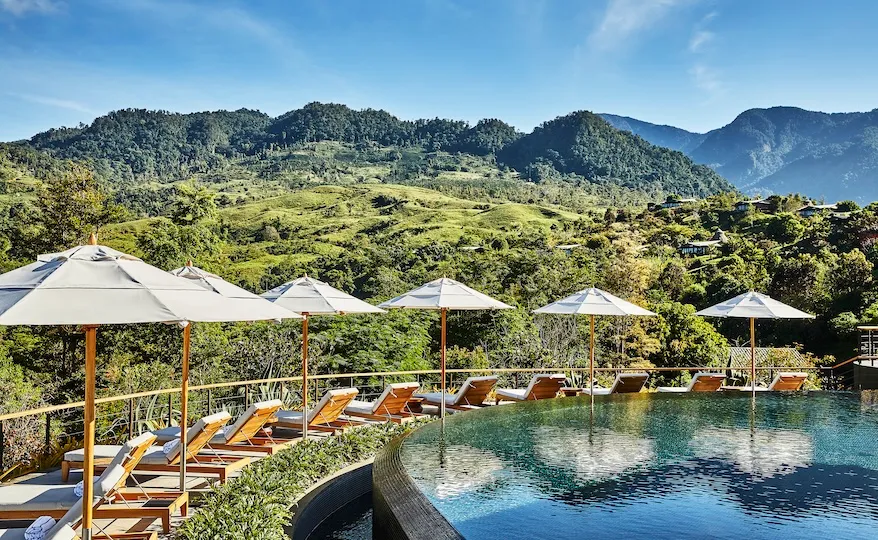 Piscina curva con tumbonas y sombrillas, con vistas a verdes colinas y montañas bajo un cielo azul despejado: un idílico paraíso de recuperación para los viajeros médicos que buscan retiros de bienestar en Costa Rica.