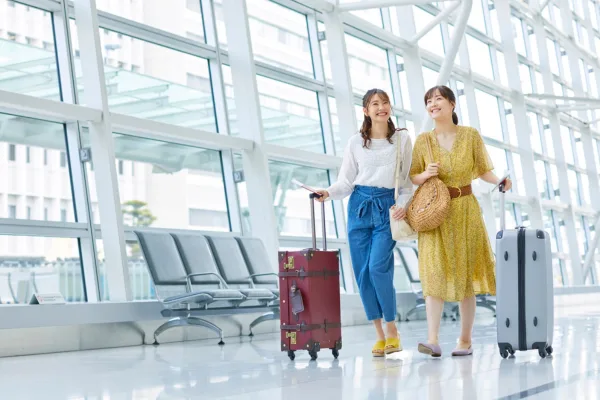 Two women walk through an airport terminal with rolling suitcases, likely discussing medical tourism packages. One wears a white top and jeans, and the other a yellow dress. They appear to be smiling and conversing.