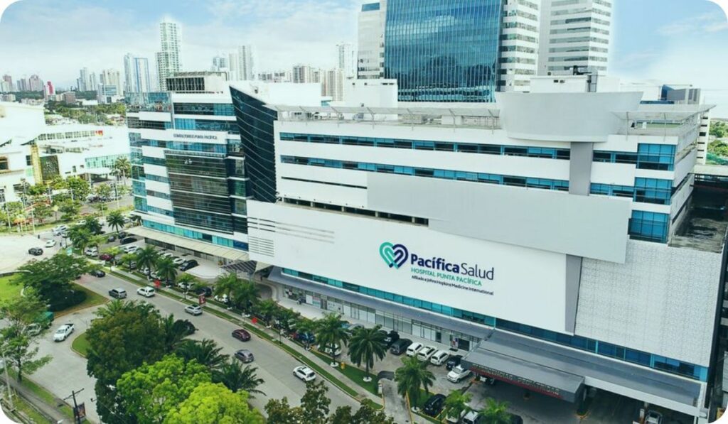 A modern multi-story hospital building with a large "Pacifica Salud" sign, surrounded by palm trees and parked cars, set in an urban area with high-rise buildings—an ideal destination for medical tourism in Panama.