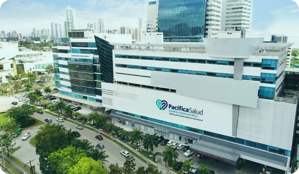 Un moderno edificio hospitalario de varias plantas con un gran letrero de "Pacifica Salud", rodeado de palmeras y coches aparcados, situado en una zona urbana con edificios altos: un destino ideal para el turismo médico en Panamá.