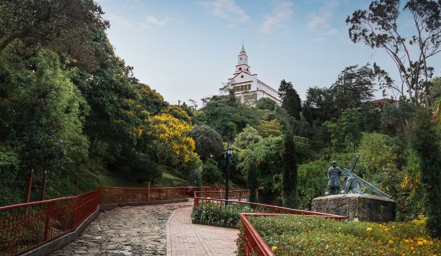 A paved pathway with red railings leads through lush greenery to a white church on a hill—a serene escape where wellness and balance are evoked by the statue and sundial on the right side of the path.