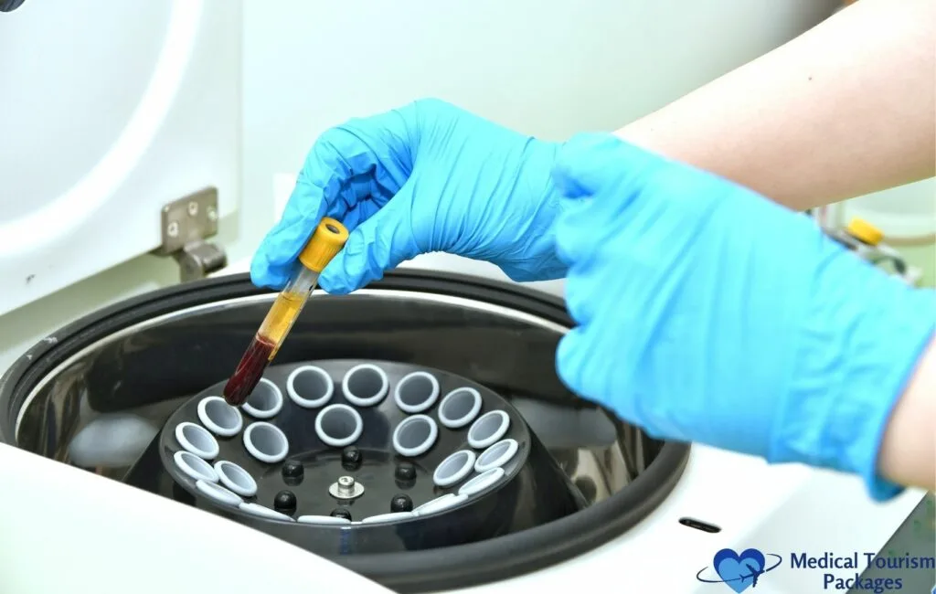 Close-up of gloved hands in bright blue medical gloves carefully placing a blood sample tube containing dark red liquid into a laboratory centrifuge with multiple circular tube holders.