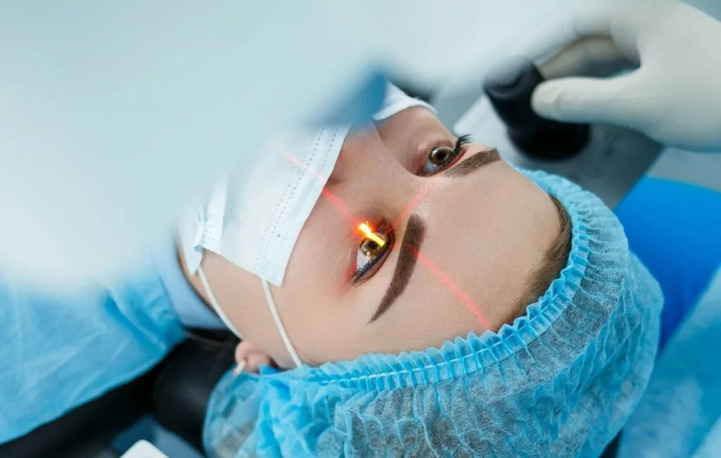 A person, possibly a medical visitor, is undergoing eye surgery with a laser being used. The individual wears a surgical cap and mask while the surgeon's gloved hand adjusts equipment.