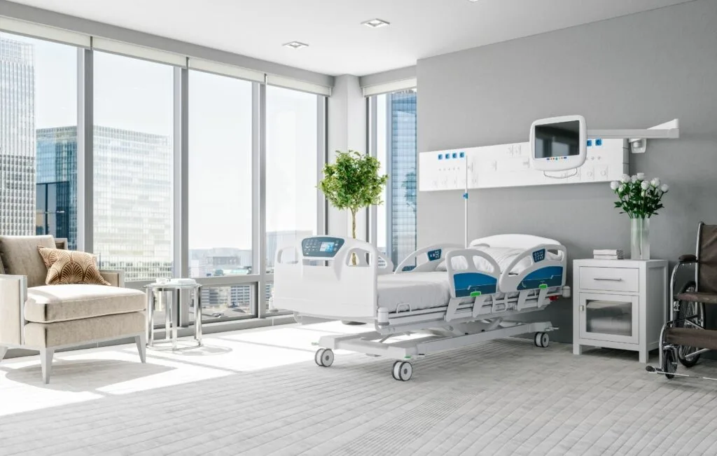 Una moderna habitación de hospital en Colombia cuenta con una cama individual, equipos médicos, una silla y grandes ventanales que revelan un paisaje urbano. Ideal para huéspedes de turismo médico que buscan un confort similar al de los hoteles, garantiza tranquilidad y una atención excepcional en medio de la bulliciosa vida urbana.