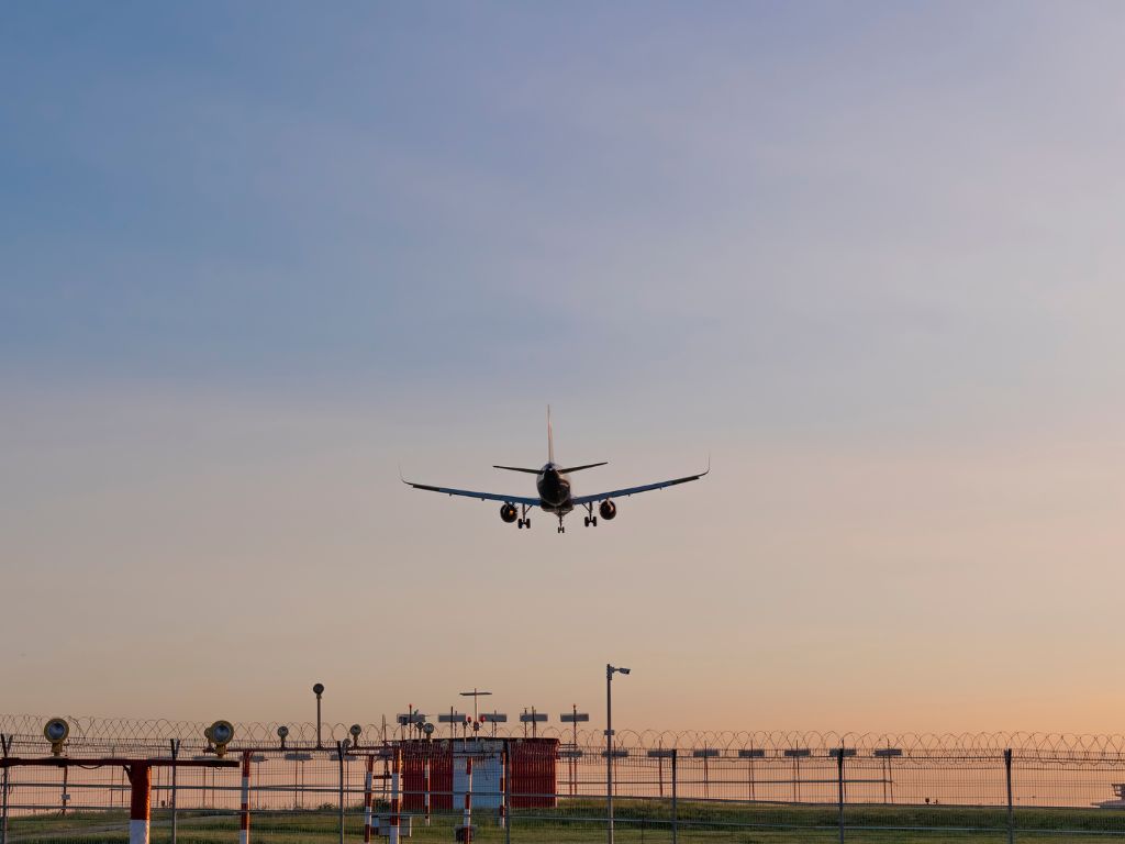 A commercial airplane is approaching a runway for landing at an airport during sunset, offering a sense of escape as airport lights and fencing glow in the foreground.