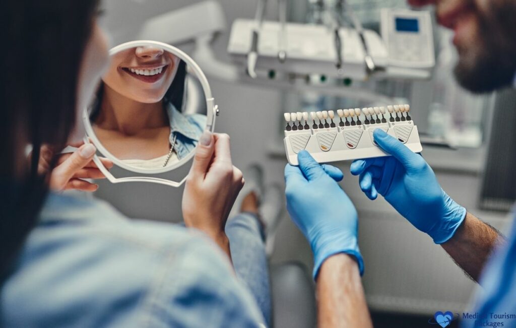 A woman sits in a dental chair, smiling at her reflection in a mirror, while a dentist holds a tooth shade guide nearby—showcasing the benefits of affordable dental care and world-class dentistry.