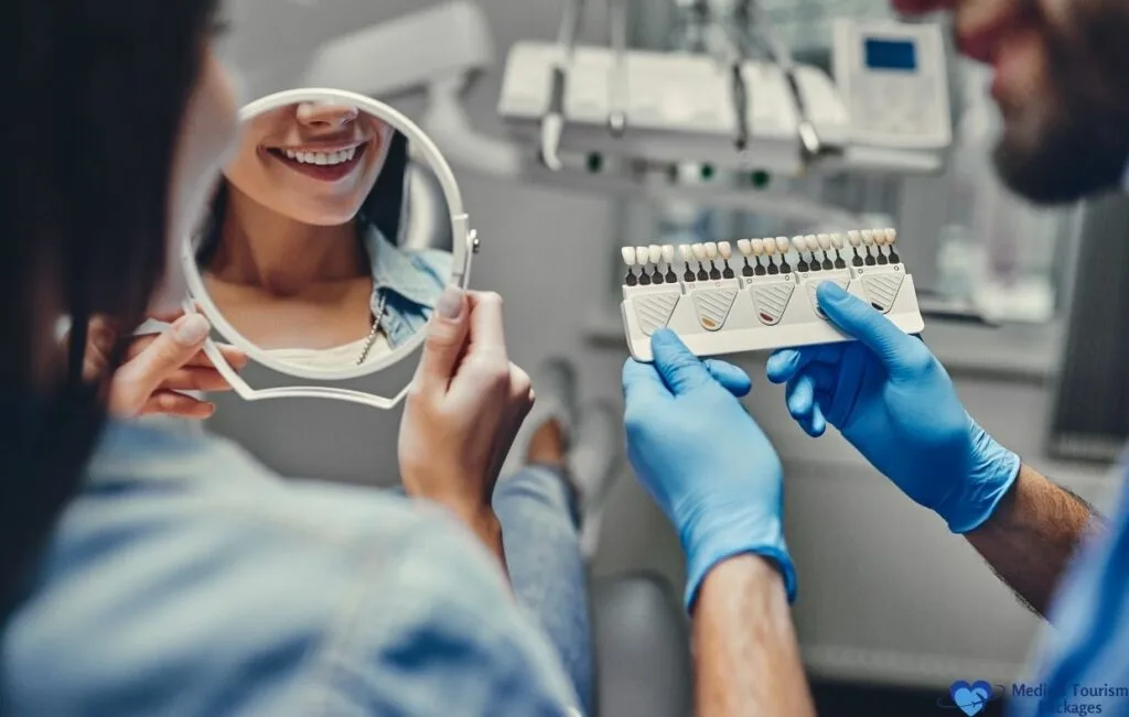 A woman sits in a dental chair, smiling at her reflection in a mirror, while a dentist holds a tooth shade guide nearby—showcasing the benefits of affordable dental care and world-class dentistry.