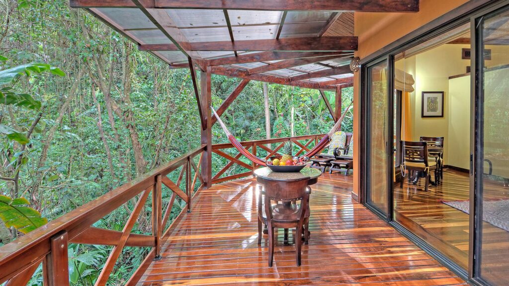 Balcón de madera con una hamaca, una mesita y sillas, rodeado de un frondoso bosque en Costa Rica, conectado a una habitación con puertas correderas de cristal-perfecto para retiros de bienestar o relajantes viajes médicos.
