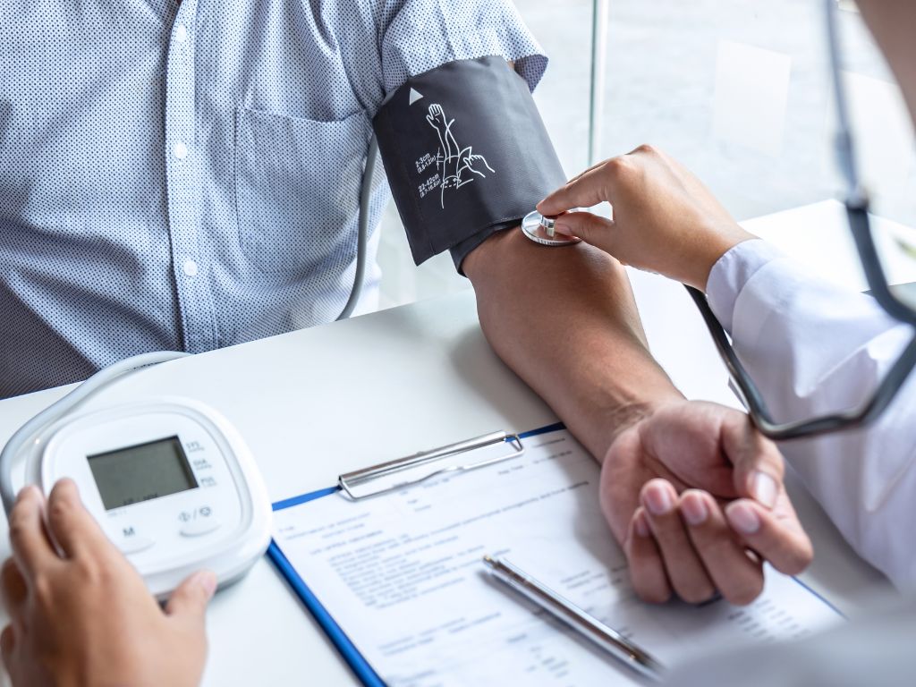 A doctor measures a patient’s blood pressure with a cuff and stethoscope, focusing on safeguarding health. A medical form and digital monitor rest on the table, supporting this preventative health check.