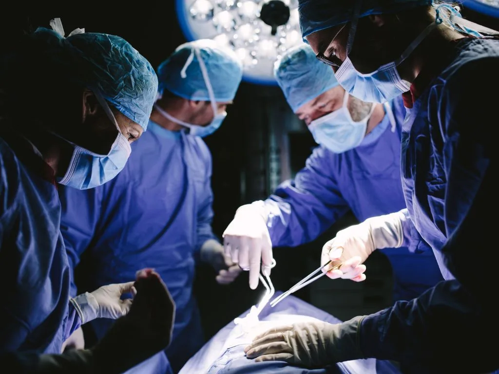 Four surgeons in surgical attire perform an operation under bright, calm lights in an operating room, focusing intently on the patient.