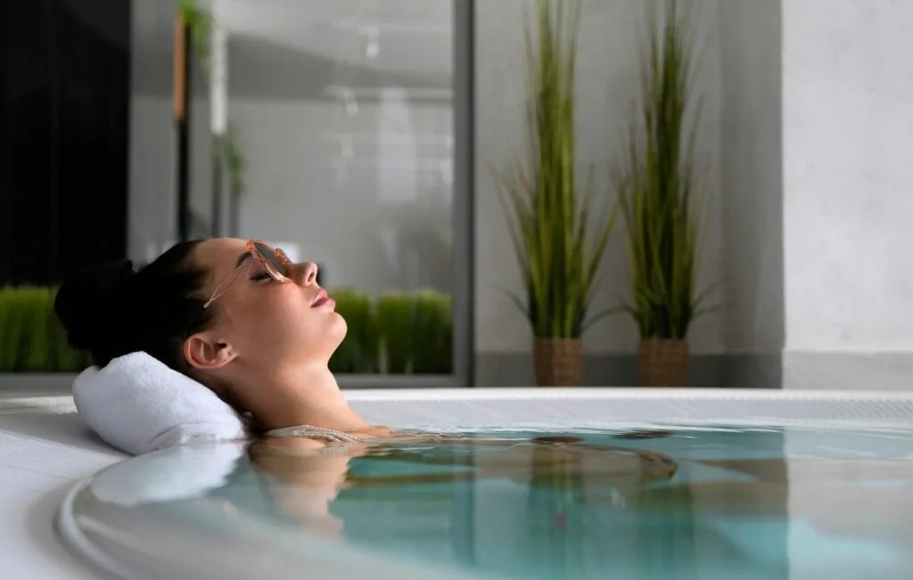 A woman with a towel under her head relaxes in a spa pool during a wellness retreat, wearing a face mask and cucumber slices on her eyes, surrounded by potted plants for ultimate stress relief.