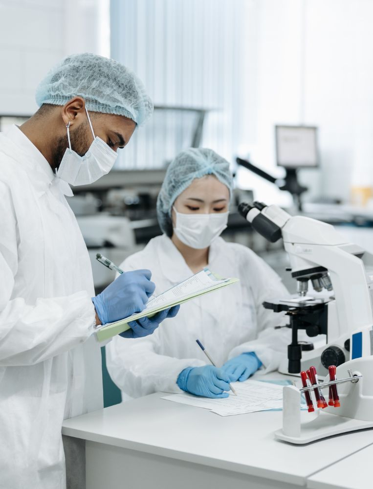 rheumathology Two scientists in lab coats, gloves, masks, and hair covers work at a laboratory table with a microscope and documents, recording observations to improve quality of life for patients undergoing rheumatology surgeries.