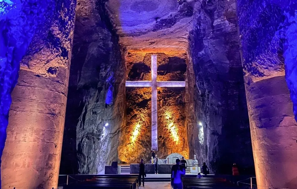 The underground salt cathedral, a marvel of Colombian culture, boasts a large illuminated cross on the stone wall. Blue and orange lighting enhances the cavernous space as visitors engage in low-impact activities, quietly observing the serene scene.