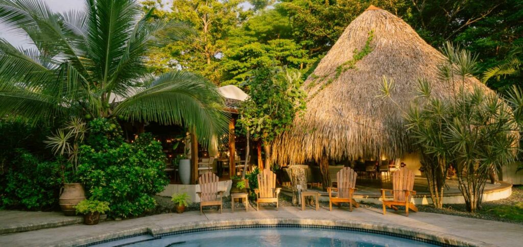 Edificio con tejado de paja rodeado de exuberante vegetación, con sillas de madera dispuestas junto a una piscina circular en primer plano, ideal para retiros de bienestar y yoga de lujo Panamá 2025.