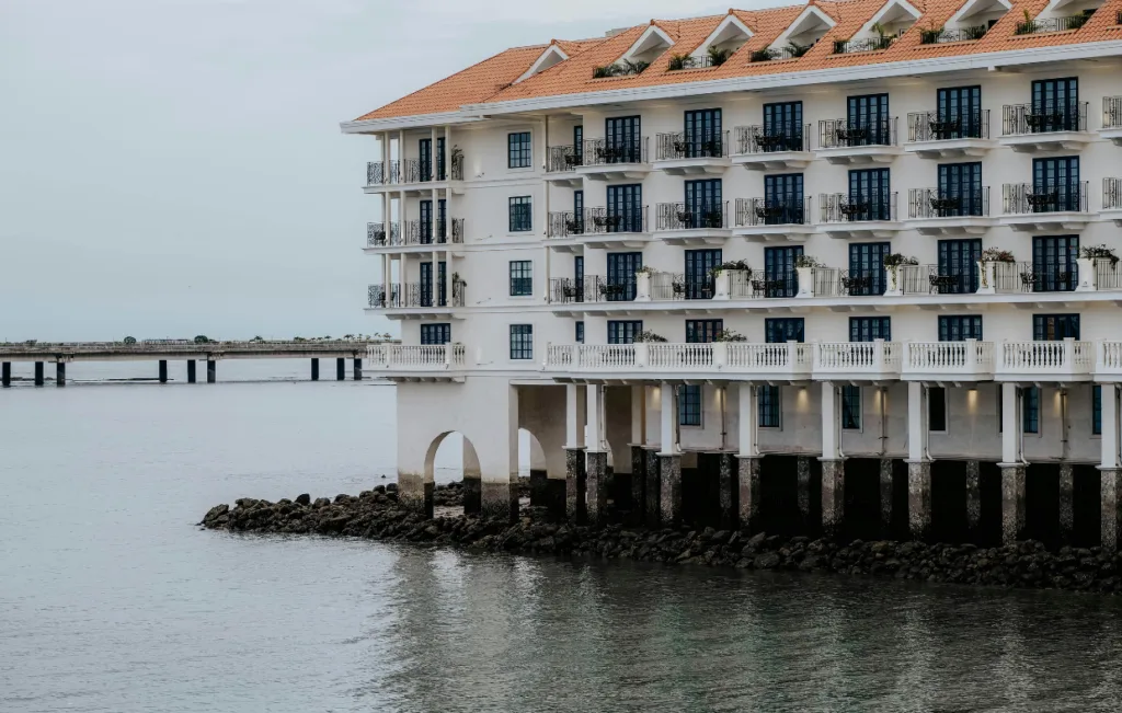 A large, multi-story building with a terracotta roof is built on stilts over water, embodying the spirit of advanced medicine. Balconies grace each level, and a distant bridge is visible in the background.