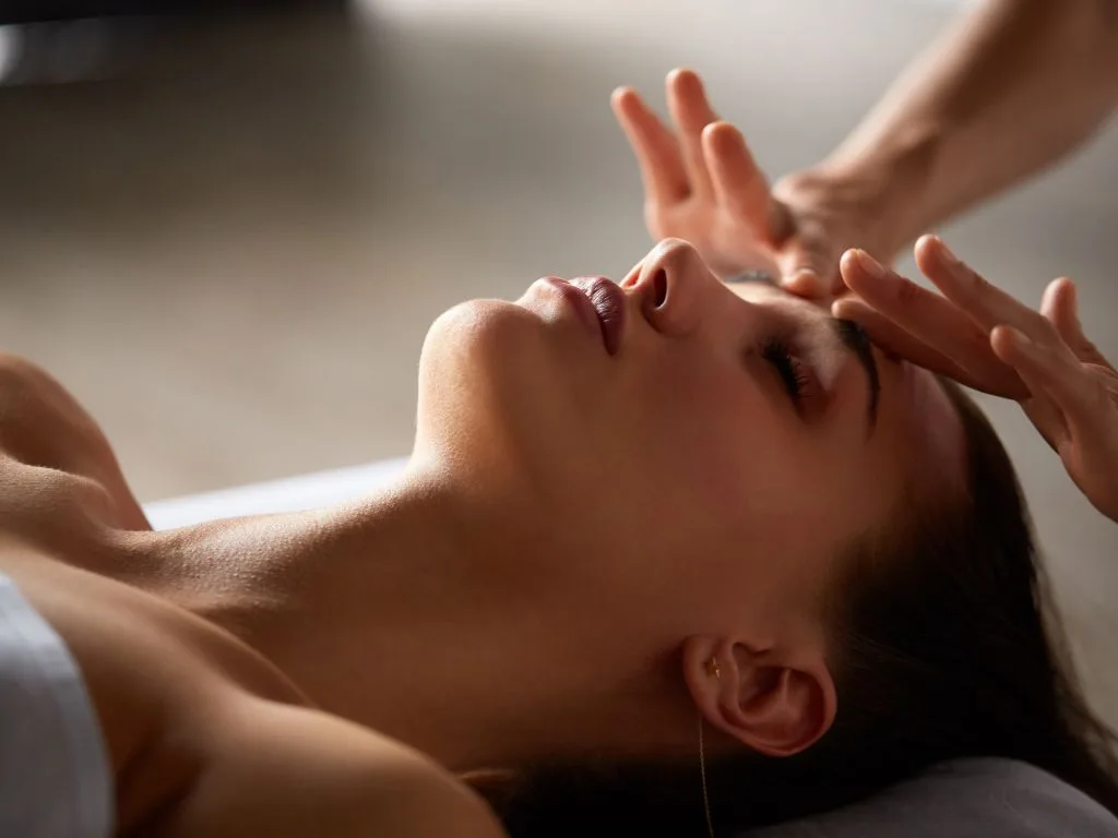 A person lies with eyes closed while another’s hands gently touch their forehead, suggesting a calm and luxurious facial massage or relaxation treatment.