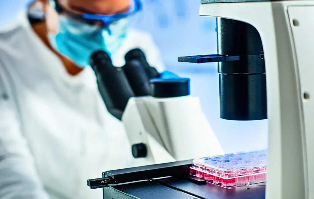 A scientist wearing a mask examines a sample through a microscope, with a multi-well plate containing pink liquid—a testament to advanced medicine—visible nearby.