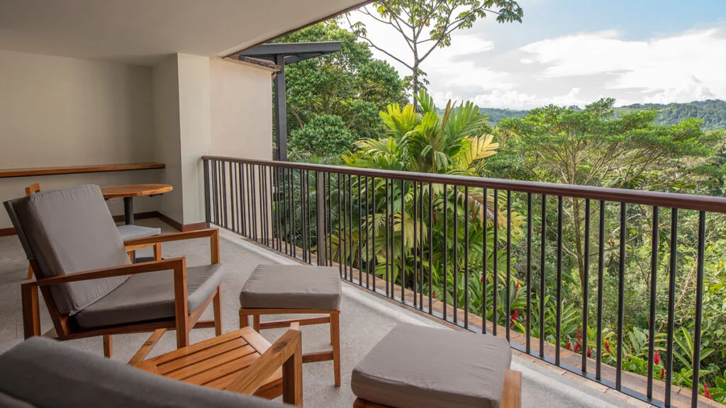 A balcony with cushioned chairs and a table overlooks lush green trees and distant hills under a partly cloudy sky—an ideal setting for medical travelers seeking a recovery paradise in Costa Rica wellness retreats.