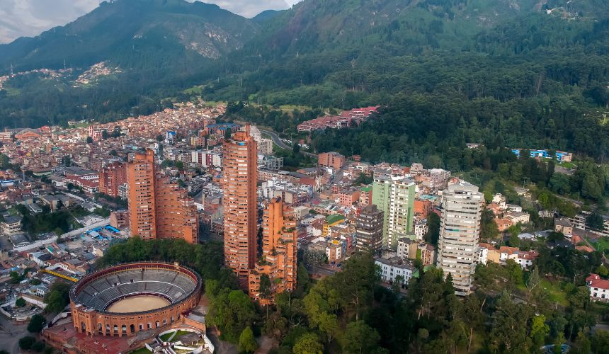 Aerial view of a city with tall buildings, a circular arena, and densely wooded hills in the background, reflecting the vitality and energy essential for safeguarding health in urban environments.