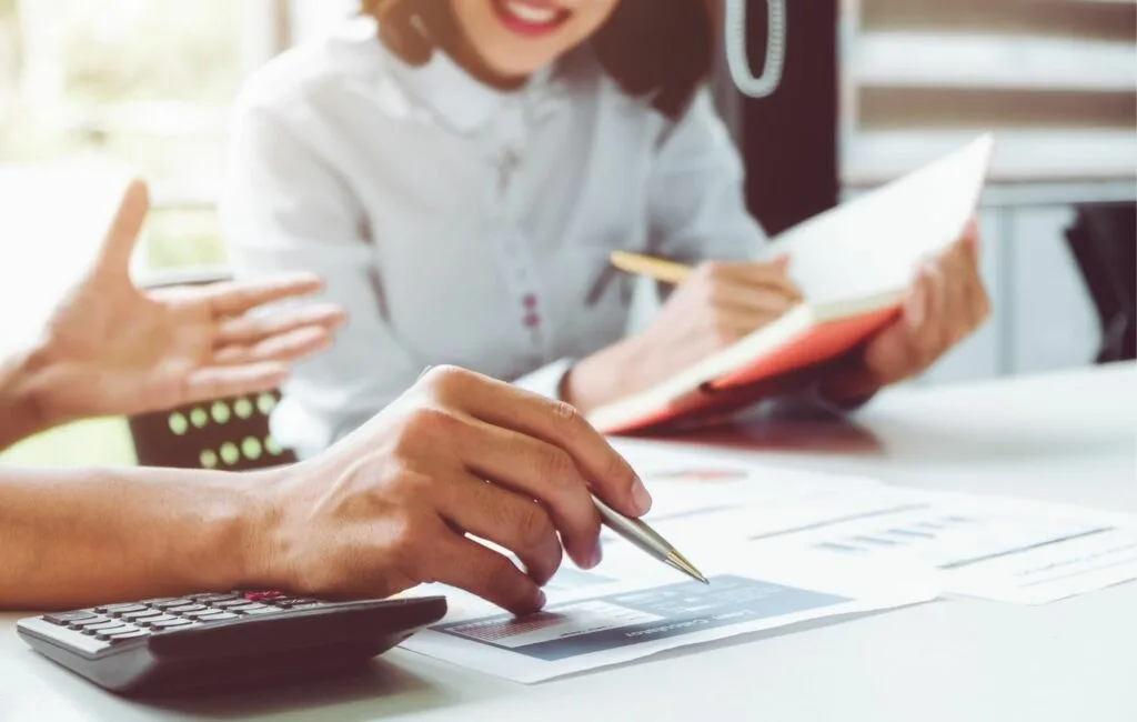 Two people sit at a desk discussing financial documents; one holds a pen and points at a paper, while the other writes in a notebook. A calculator is also visible, suggesting they may be reviewing costs for premium care from top doctors or plastic surgeons Colombia.