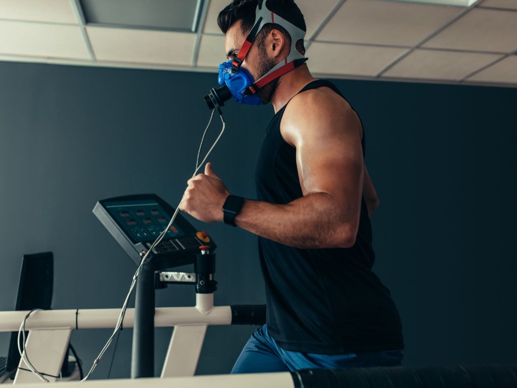 A man runs on a treadmill wearing a high-tech mask and connecting wires, likely undergoing a preventative health or respiratory test in a gym or lab setting to assess and safeguard his vitality.