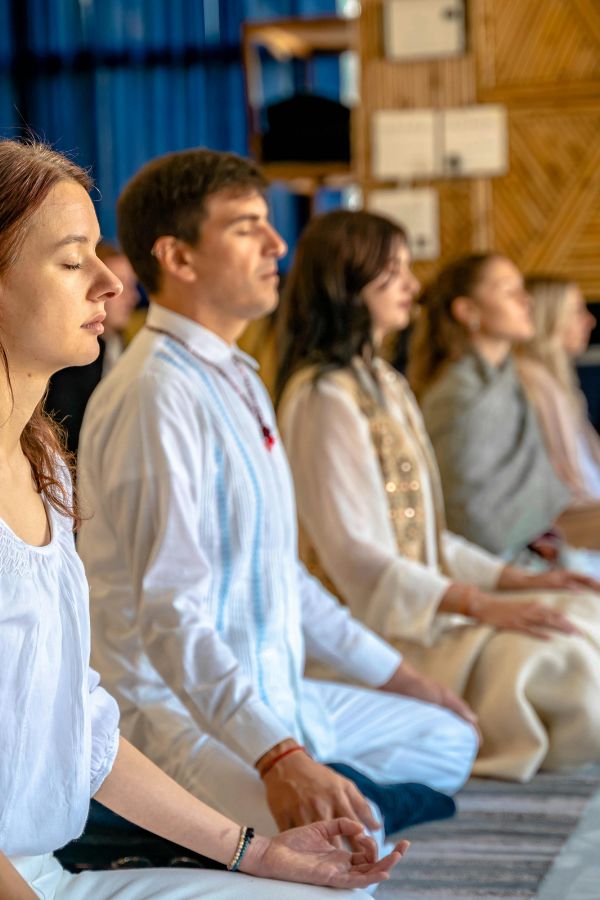 A group of people sit cross-legged on the floor with eyes closed, meditating in a calm indoor setting, finding balance and wellness together.