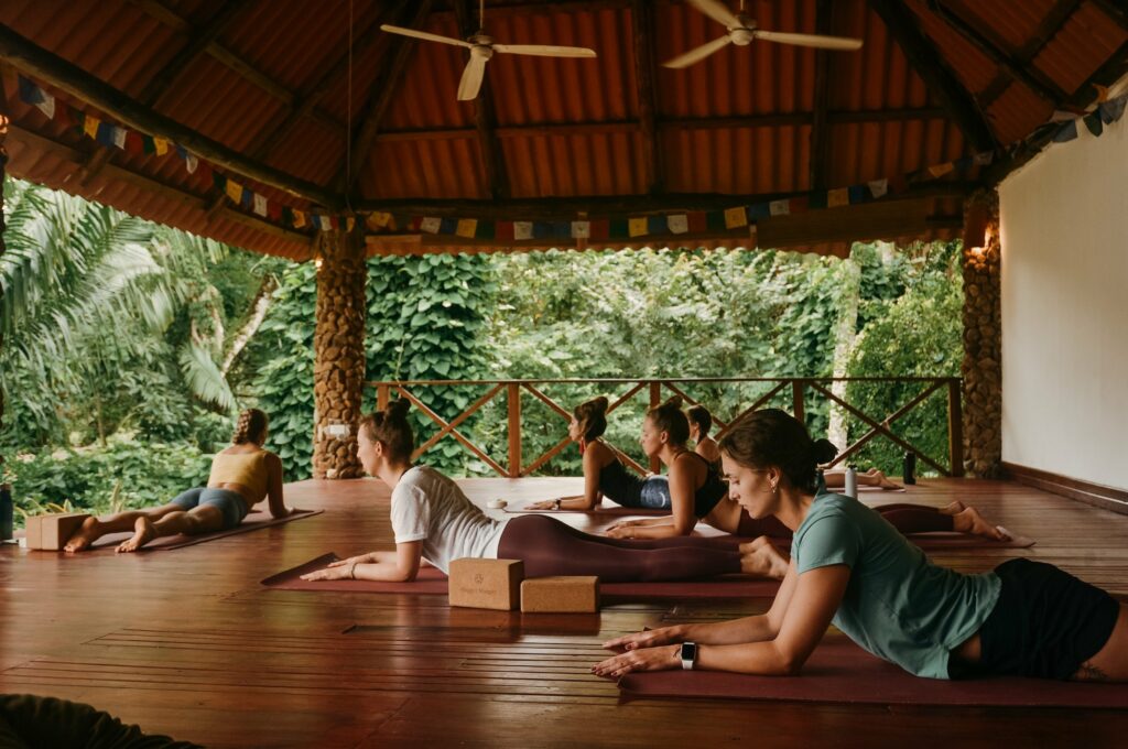 Un grupo de personas practica yoga sobre esterillas en un pabellón cubierto de madera rodeado de exuberante vegetación.