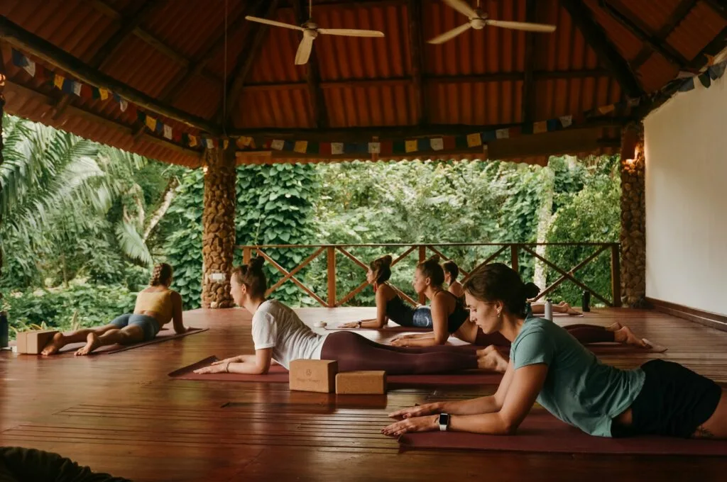 Un grupo de personas practica yoga sobre esterillas en un pabellón cubierto de madera rodeado de exuberante vegetación.