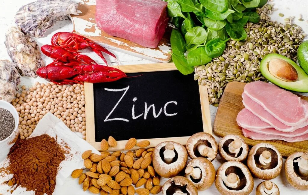 Alrededor de una pizarra rotulada "Zinc", una abundancia de alimentos ricos en zinc -carne, marisco, semillas, espinacas, aguacates, almendras, setas y lentejas- muestra los elementos esenciales para una nutrición y un bienestar óptimos.